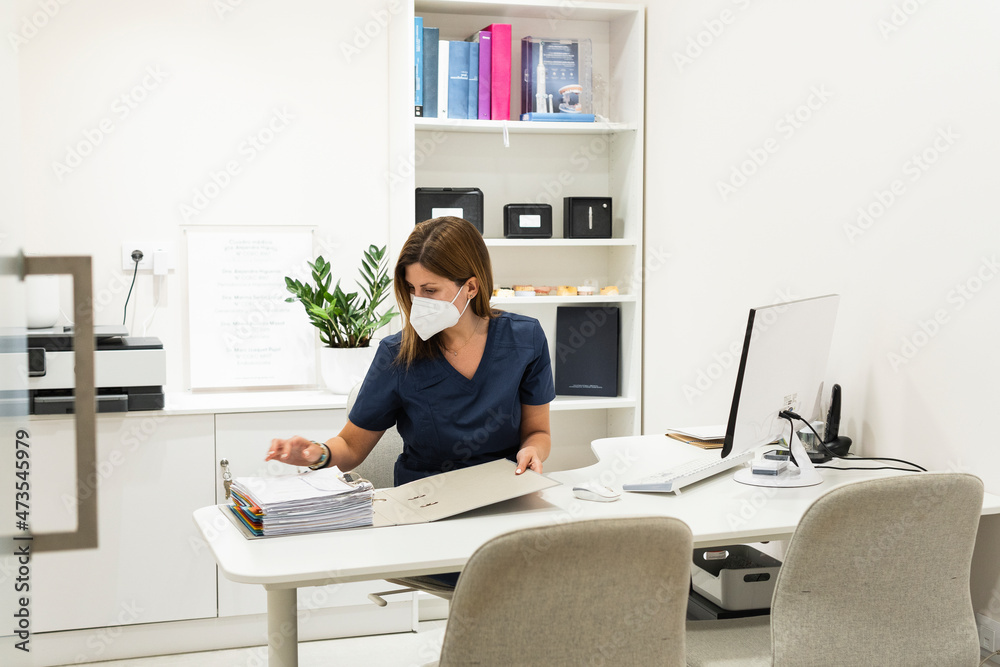 Female dentist wearing protective face mask checking medical records at desk in clinic Female dentist wearing protective face mask checking medical records at desk in clinic
