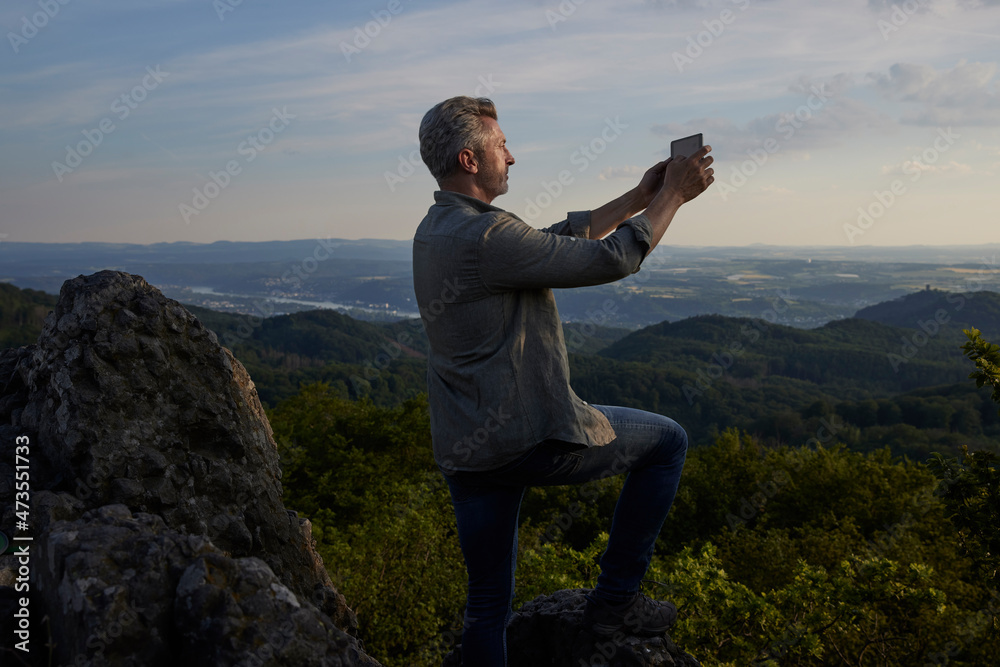Male hiker taking photo through digital tablet on mountain during sunset Male hiker taking photo through digital tablet on mountain during sunset