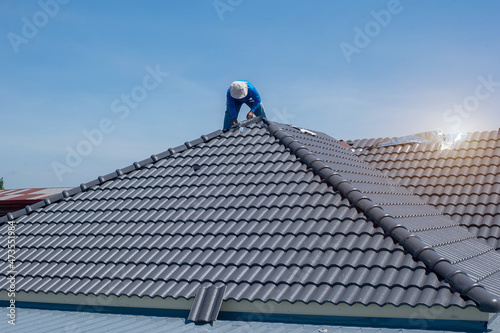 Roof repair, worker with white gloves replacing gray tiles or shingles on house with blue sky as background and copy space, Roofing - construction worker standing on a roof covering it with tiles.
