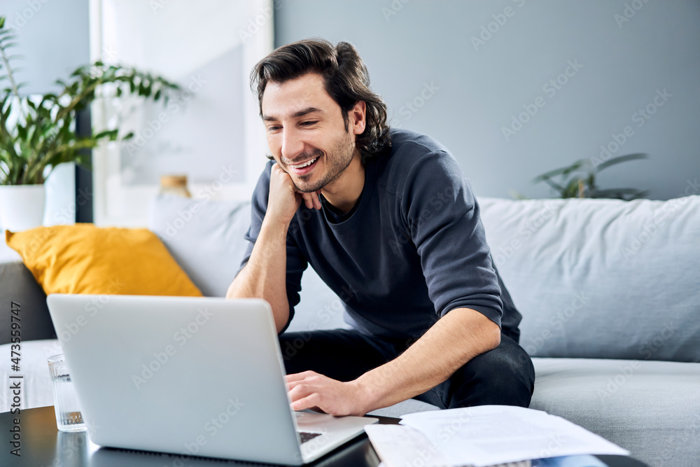 Smiling businessman with hand on chin using laptop at home Smiling businessman with hand on chin using laptop at home