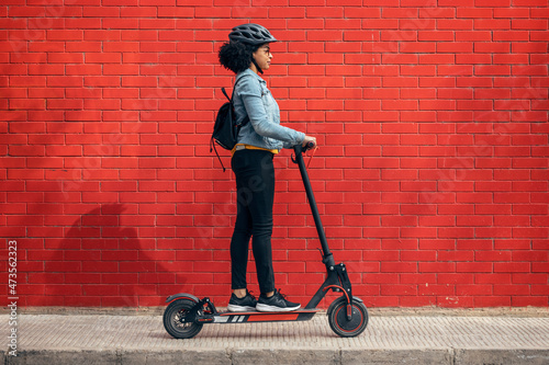 Young woman with backpack riding electric push scooter by red wall photo