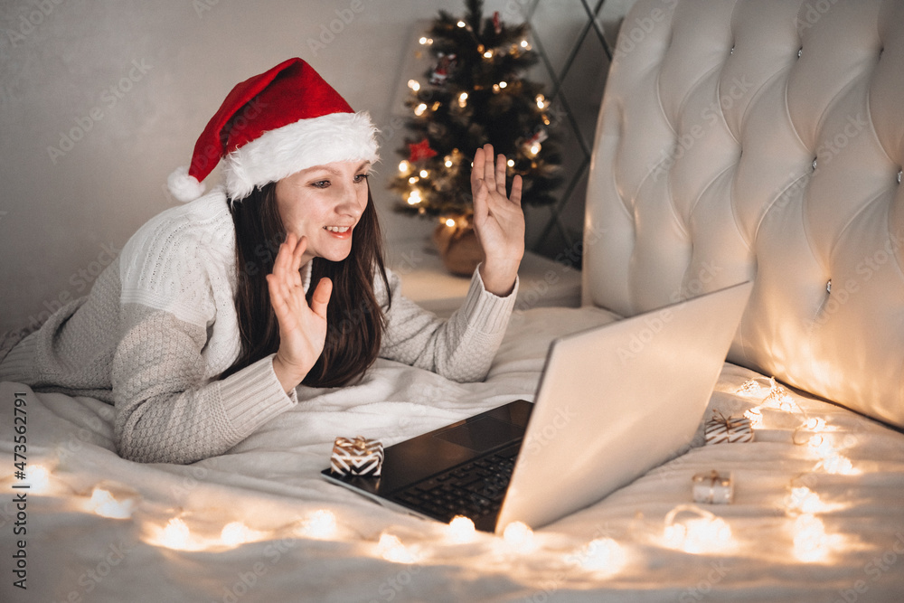 Young woman in red santa hat having video chat with family and friend. Smiling brunette girl talking online video call lying on bed. Christmas and new year holiday. Social media and internet Young woman in red santa hat having video chat with family and friend. Smiling brunette girl talking online video call lying on bed. Christmas and new year holiday. Social media and internet