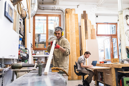 Mature male carpenter working on wood while coworker writing in workshop photo