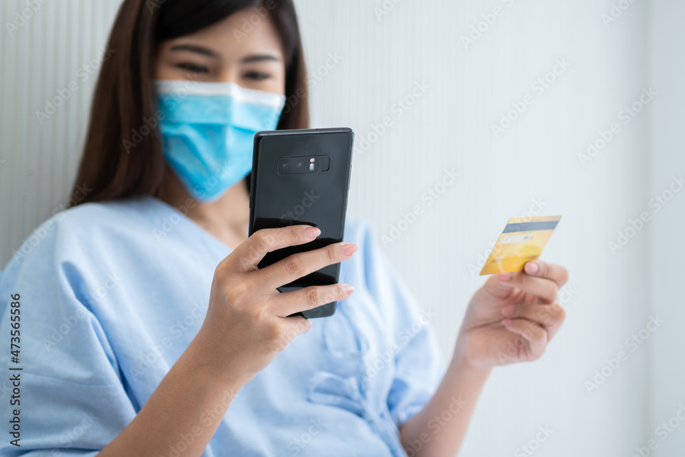 Happy Asian woman wearing a medical mask and holding mock up credit /insurance card and smartphone in a hospital bed for Check health insurance eligibility. Insurance policy by bank, payment medical Happy Asian woman wearing a medical mask and holding mock up credit /insurance card and smartphone in a hospital bed for Check health insurance eligibility. Insurance policy by bank, payment medical
