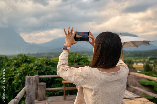 Asian woman taking a video of mountain view by smartphone on wooden balcony in vacation at evening