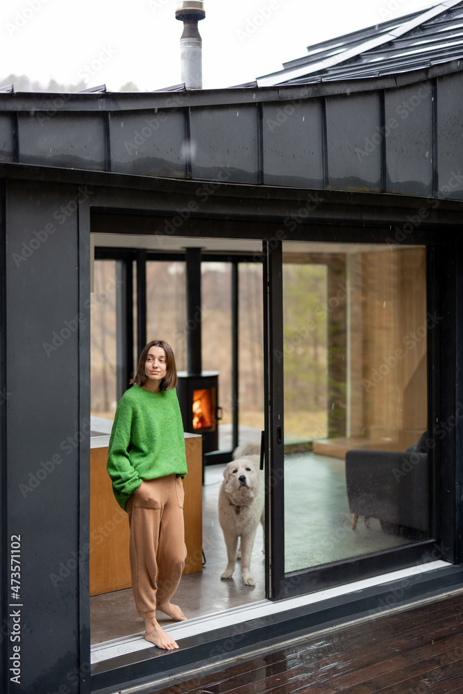 Young woman with her dog at modern house on nature. Girl in green sweater standing near the window and look outside. Concept of comfort and happy modern life Young woman with her dog at modern house on nature. Girl in green sweater standing near the window and look outside. Concept of comfort and happy modern life