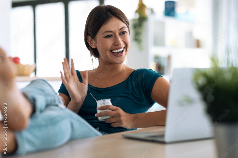 Confident young woman having a videocall with computer while while eating yogurt sitting in the kitchen at home.