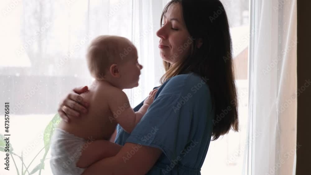 Beautiful woman holds a newborn baby. Loving Mother hugging of her baby at home. Bright portrait of happy mum holding child on hands. Loving mommy staying for a window and soothing adorable girl. Beautiful woman holds a newborn baby. Loving Mother hugging of her baby at home. Bright portrait of happy mum holding child on hands. Loving mommy staying for a window and soothing adorable girl.