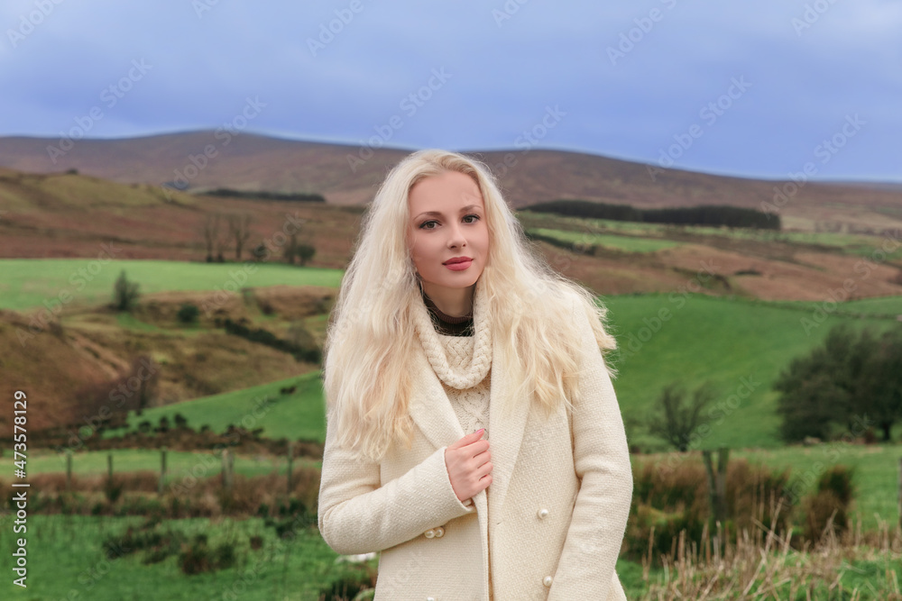 Beautiful young caucasian blonde woman in a white jumper standing in the field overlooking picturesque mountains. Beautiful young caucasian blonde woman in a white jumper standing in the field overlooking picturesque mountains.