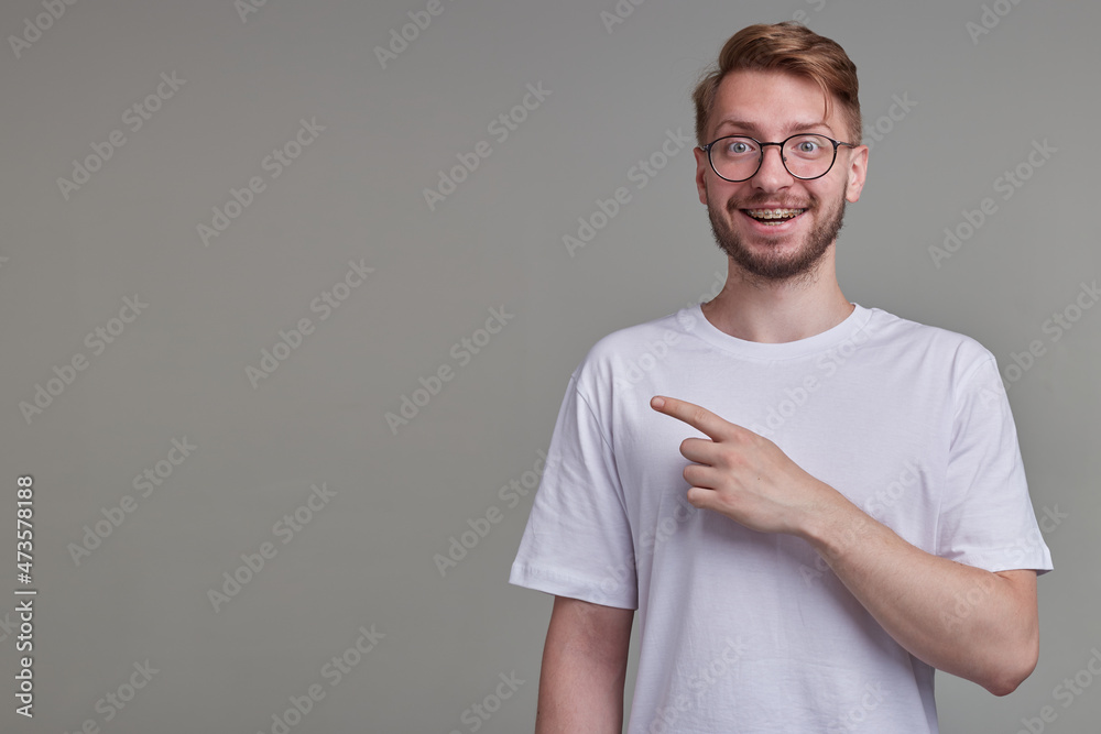 studio portrait of young bearded man wears white t-shirt point with a finger aside at copy space, smiles broadly and feels happy isolated over gray background. studio portrait of young bearded man wears white t-shirt point with a finger aside at copy space, smiles broadly and feels happy isolated over gray background.