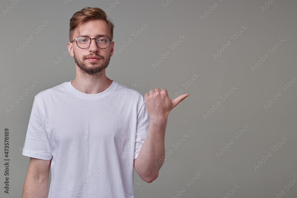 studio portrait of young bearded man wears white t-shirt and glasses pointing aside with a finger with serious facial expression, isolated over gray background. studio portrait of young bearded man wears white t-shirt and glasses pointing aside with a finger with serious facial expression, isolated over gray background.