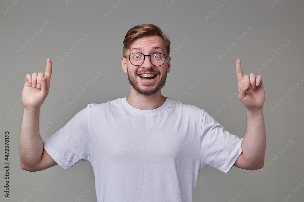 Studio portrait of young bearded man, wears casually posing over gray studio background points with a finger upwards, smiles broadly with happy, shocked facial expression. isolated. Studio portrait of young bearded man, wears casually posing over gray studio background points with a finger upwards, smiles broadly with happy, shocked facial expression. isolated.