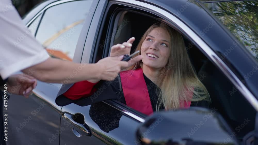 Charming slim woman in graduation gown sitting in car as unrecognizable parent passing key in slow motion. Portrait of surprised happy beautiful Caucasian grad receiving vehicle as gift smiling