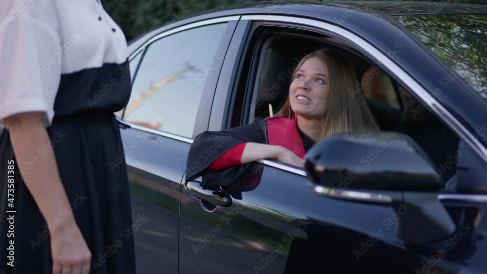 Young laughing graduate woman sitting in car talking with unrecognizable parent outdoors. Portrait of excited happy Caucasian daughter chatting with mother on graduation day leaving for ceremony Young laughing graduate woman sitting in car talking with unrecognizable parent outdoors. Portrait of excited happy Caucasian daughter chatting with mother on graduation day leaving for ceremony