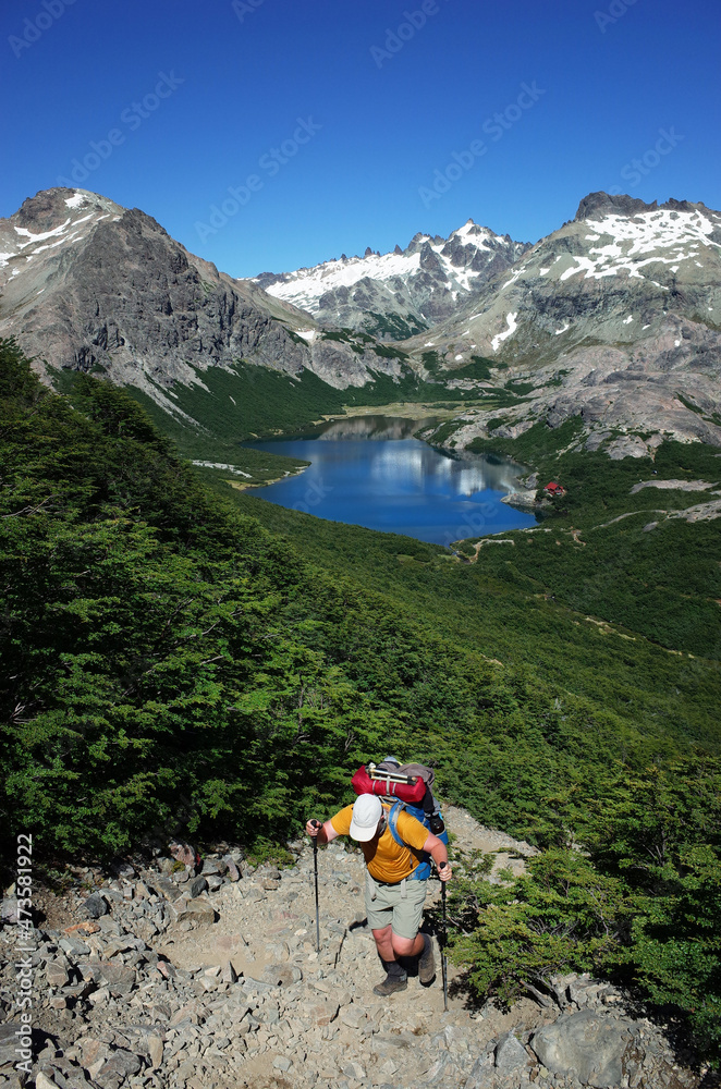 Trekking in Patagonia, Picturesque mountain lake, Man with big backpack hiking uphill on gray stony path in green forest with view of Jakob lagoon in Nahuel Huapi National Park, Argentina