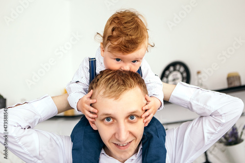 Little boy cute child redhead baby sitting on neck, father playing with his son. Toddler having fun at home dressed overalls suspenders and white shirt. Happy childhood, family concept