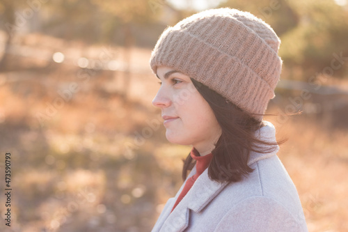 A serious girl in a knitted hat stands against a background and a coat against the backdrop of an autumn pale park landscape.