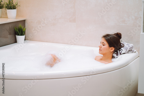 Young woman lies in white bath of foam with her eyes closed. Spa procedures, cleansing, relaxation, self-care.