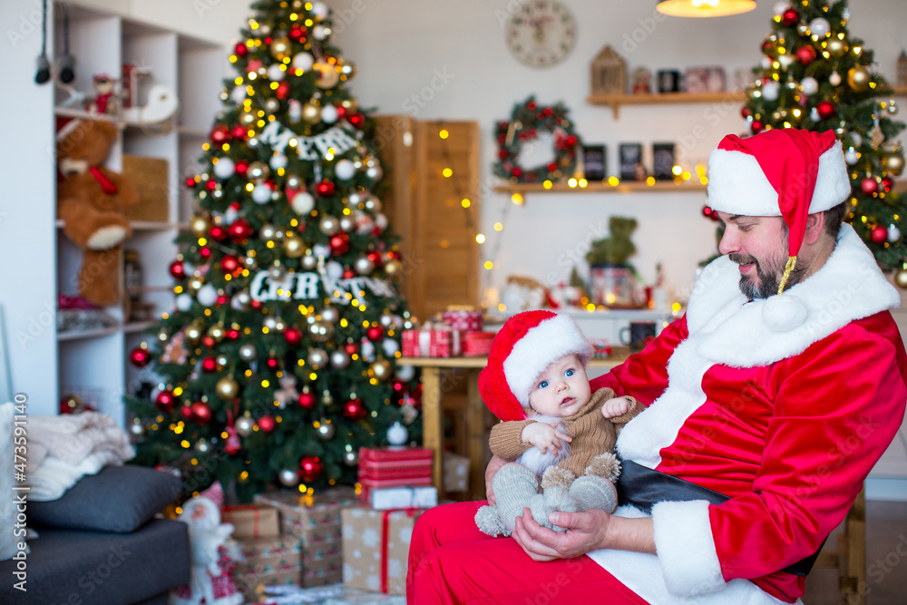 Big and small Santa Claus are sitting against the background of Christmas trees decorated with balls and garlands Big and small Santa Claus are sitting against the background of Christmas trees decorated with balls and garlands