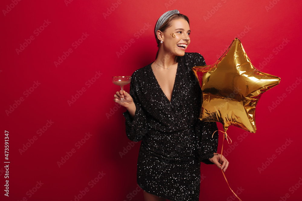 Funny young fair-skinned woman looking to side, celebrating christmas in chic dress on red background. Girl model is having fun, drinking wine from glass. Holiday concept, leisure, lifestyle Funny young fair-skinned woman looking to side, celebrating christmas in chic dress on red background. Girl model is having fun, drinking wine from glass. Holiday concept, leisure, lifestyle