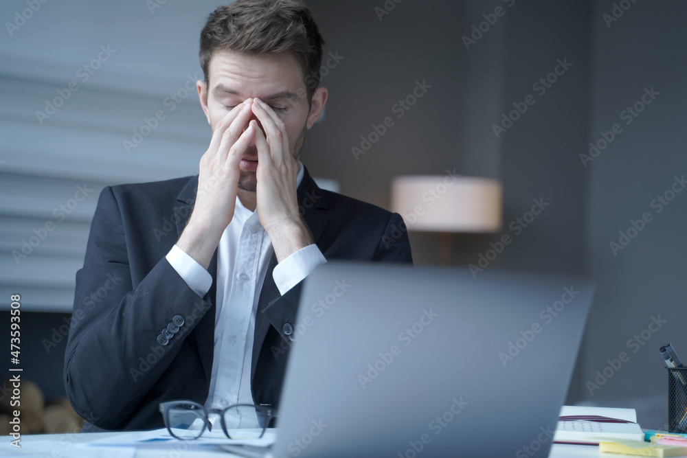Overworked male entrepreneur sits at home office, suffering from heavy headache and fatigue Overworked male entrepreneur sits at home office, suffering from heavy headache and fatigue