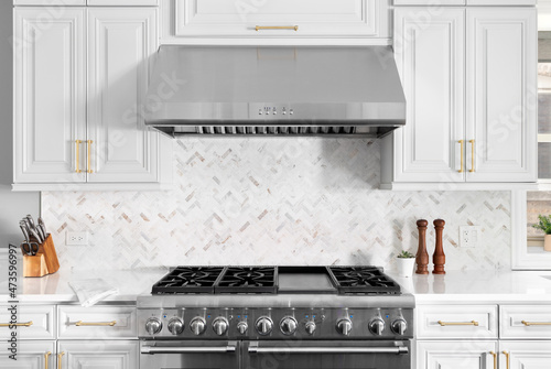 A detail shot of a beautiful white kitchen's stainless steel stove, hood, granite counter tops, and a herringbone marble tiled back splash.