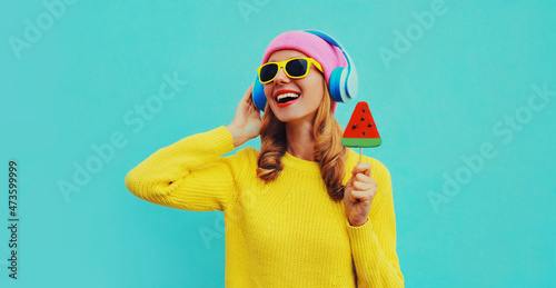 Summer fresh colorful portrait of happy laughing young woman in headphones listening to music with fruit juicy lollipop or ice cream shaped slice of watermelon on blue background