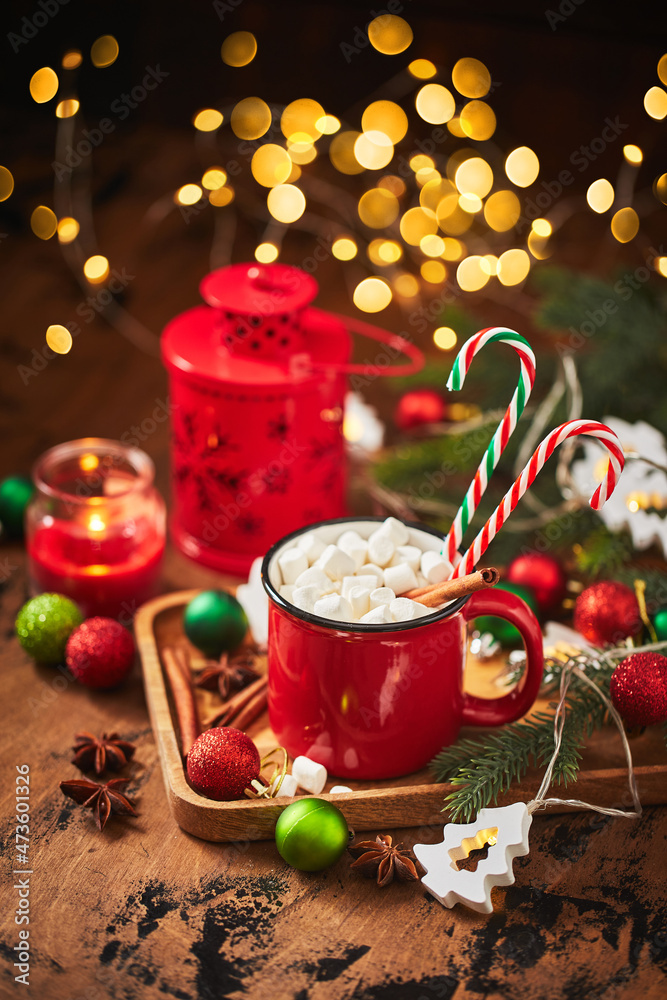 Red cup of hot cocoa with marshmallows and candy canes on wooden table with christmas tree and glowing garland for christmas