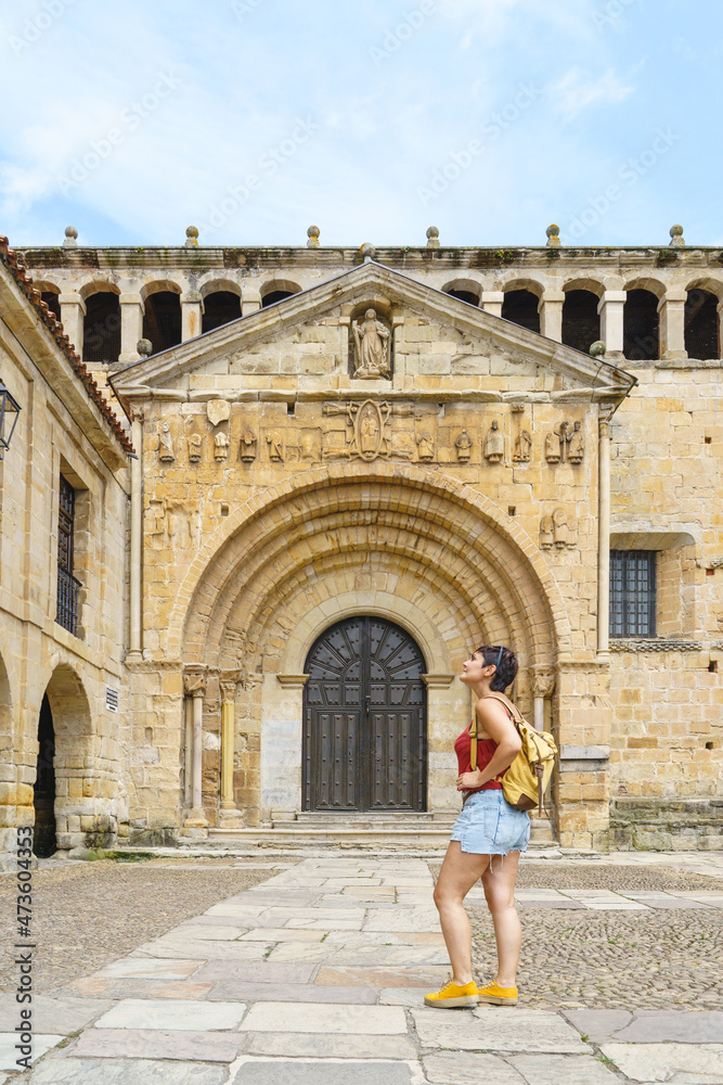 Horizontal view of young woman with backpack sightseeing. Panoramic view of woman looking at the ancient cathedral of Santillana del Mar. Travel and holidays concept in european cities.