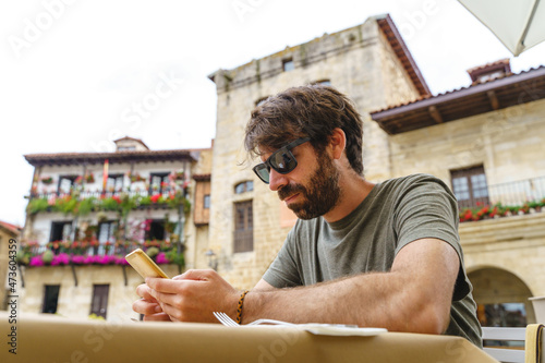 Side view of caucasian beard man looking at his phone outdoors. Horizontal view of man with sunglasses at a restaurant using technology. Technology and people concept.