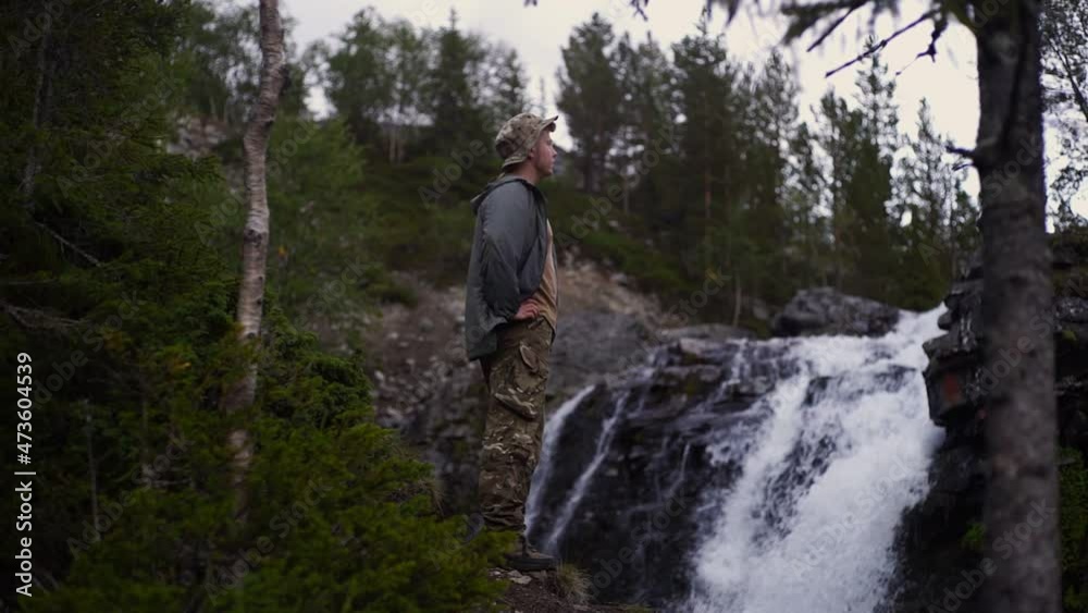 A lonely traveler stands in a heroic pose near a waterfall and admires the mountains A lonely traveler stands in a heroic pose near a waterfall and admires the mountains