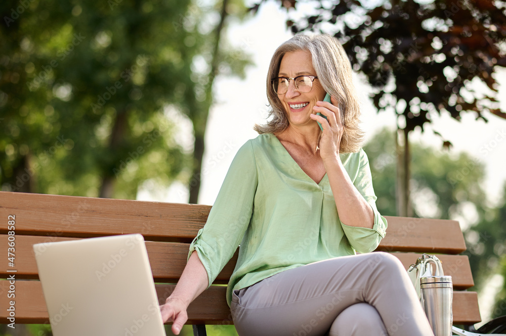 Woman working on laptop sitting with smartphone on bench Woman working on laptop sitting with smartphone on bench