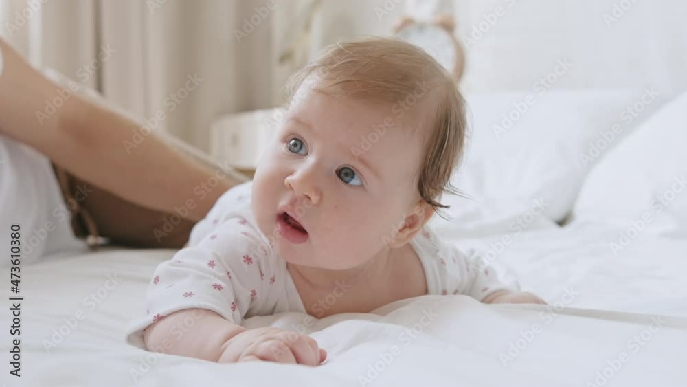 Cute sweet baby newborn with blue eyes lying on white bed sheet with parents, expressive looks around with interest and moving her lips. Third month closeup face portrait. Baby is lying on his tummy Cute sweet baby newborn with blue eyes lying on white bed sheet with parents, expressive looks around with interest and moving her lips. Third month closeup face portrait. Baby is lying on his tummy