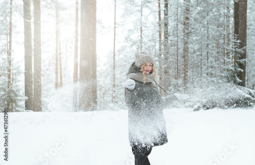 white young woman in a hat and mittens and a black jacket throwing snow in winter in the forest among the trees