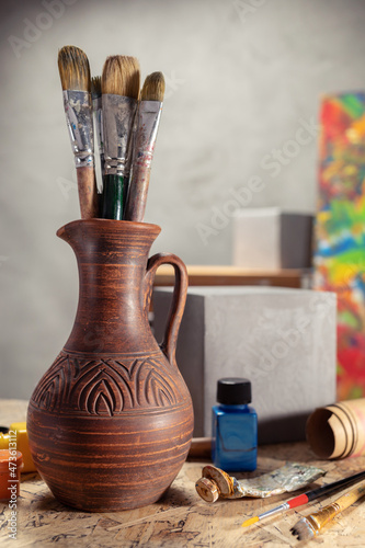 Artist supplies at wooden table. Paint brush in clay jug and art painter tool on desk background
