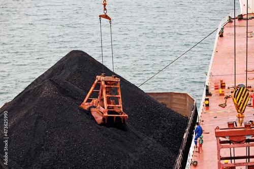 Loading coal from cargo barges onto a bulk carrier using ship cranes and grabs at the port of Muara Pantai, Indonesia. January,2021.