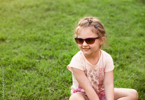 Fun excited happy kid girl in sunglasses sitting on green glass outdoor summer background in sunny bright day. Closeup © nastia1983
