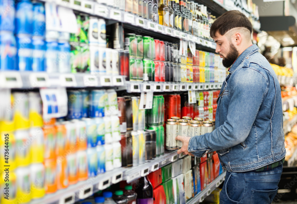 Interested young glad cheerful positive man making purchases in grocery store, buying canned beer