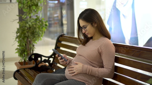 A young pregnant woman in glasses sits with a phone on a bench. Girl in the mall and uses a smartphone.