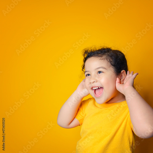 Little Asian girl holds her hand near her ear and listenings. Exciting face on Asian child girl. Exciting face on happy asian girl wear yellow shirt and listening to curious good news.