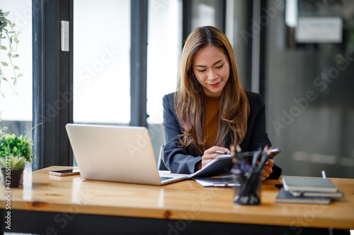 Asian young businesswoman sitting at a table with laptop computer and doing math finance report on wooden desk, tax, accounting, statistics and analytical research concept © David