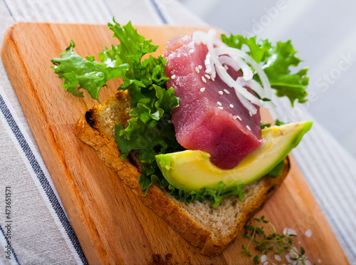Slice of grilled bread with raw tuna slice and avocado served on wooden board with fresh greens