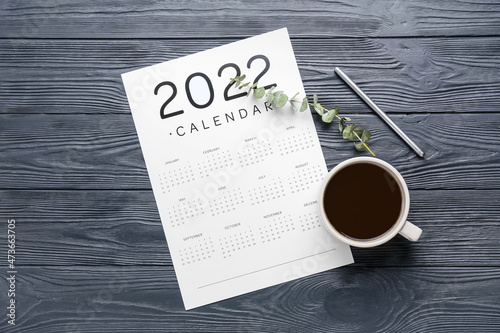 Paper calendar for 2022 year, pen, cup of coffee and eucalyptus branch on dark wooden background