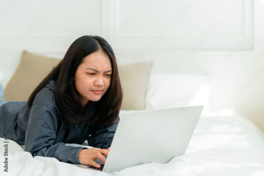 Portrait, a cute Asian teenage girl. wearing gray shirt lying in bed with a notebook computer in the bedroom to study online with a thoughtful face Portrait, a cute Asian teenage girl. wearing gray shirt lying in bed with a notebook computer in the bedroom to study online with a thoughtful face