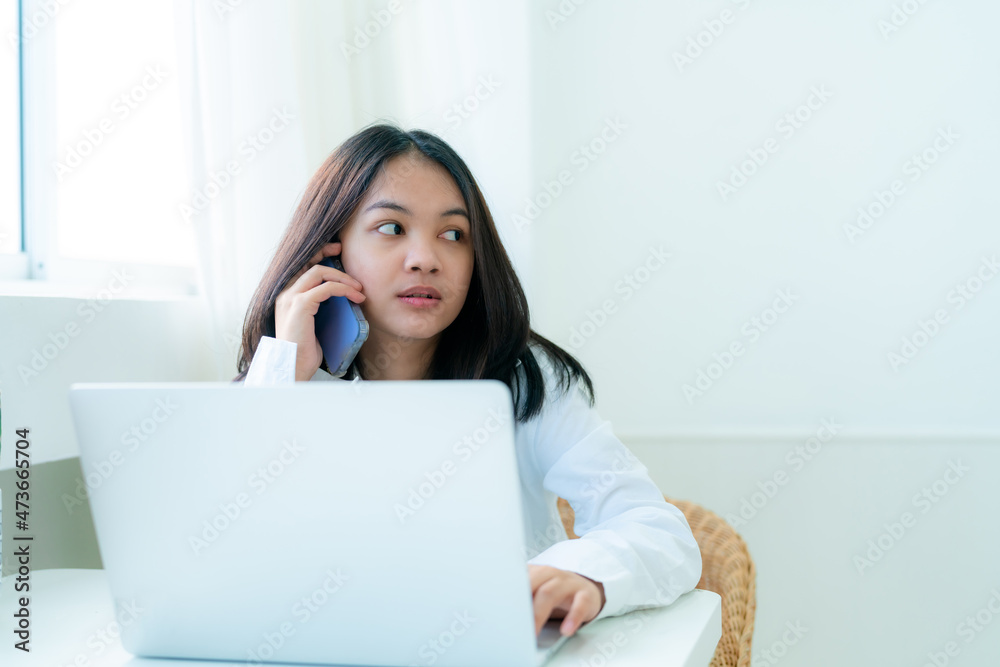 Portrait Cute Asian teen with long hair wearing a white shirt talking on the phone and study online with a laptop computer on the desk happily Portrait Cute Asian teen with long hair wearing a white shirt talking on the phone and study online with a laptop computer on the desk happily