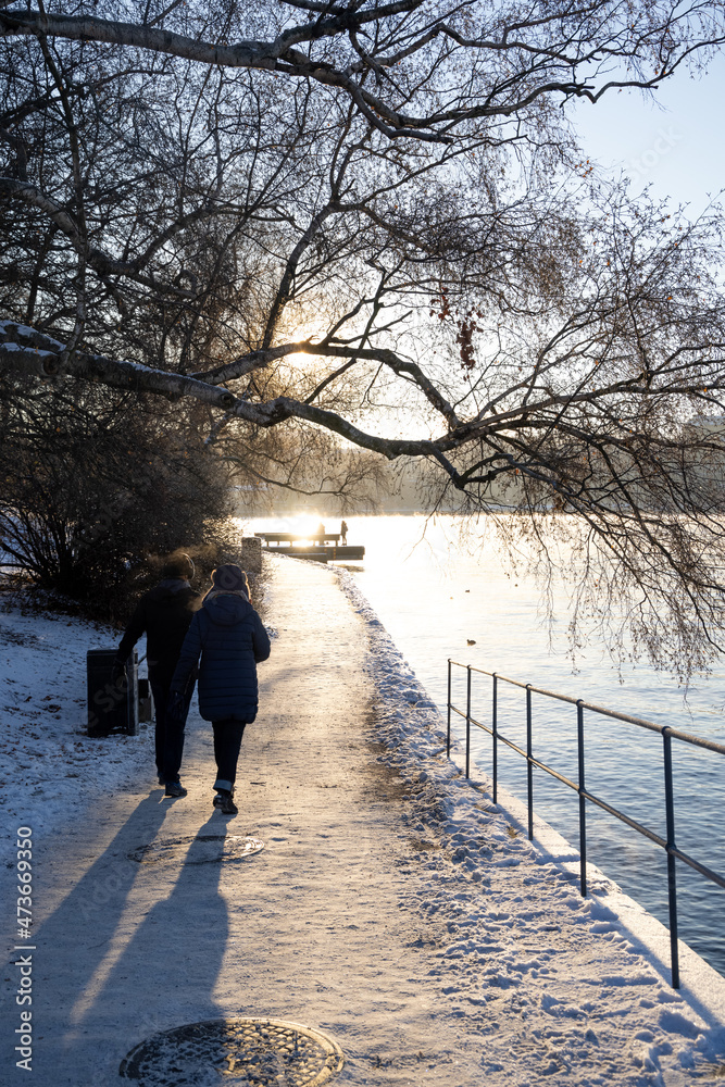 People walking on a walking path at Stockholm bay on freezing winter day with steaming water, birch tree in front and cozy sun light. Winter time in capital of Sweden  