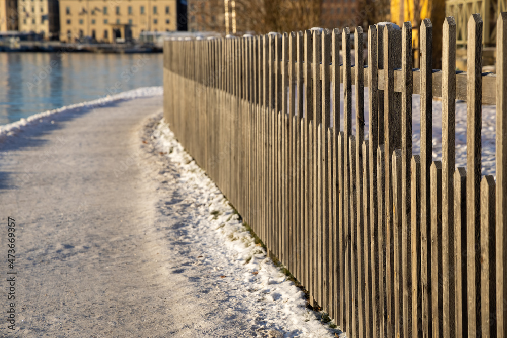 Snowy walking path at Stockholm bay on sunny freezing winter day with steaming water, wooden Scandinavian fence in front and city buildings in a background. Winter time in capital of Sweden Snowy walking path at Stockholm bay on sunny freezing winter day with steaming water, wooden Scandinavian fence in front and city buildings in a background. Winter time in capital of Sweden