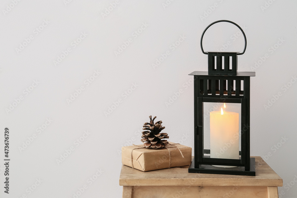 Black lantern with burning candle, gift and fir cone on stool near light wall