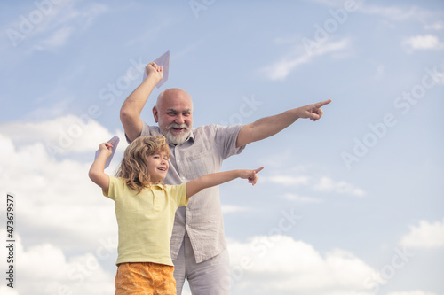 Young grandson and old grandfather playing with toy paper plane against summer sky background.