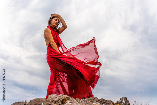 A woman in a red dress stands above a stormy sky, her dress fluttering, the fabric flying in the wind.